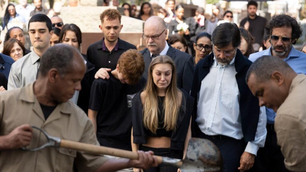 Carla Dias, como Suzane Richthofen vestida de preto chora junto ao seu irmão, familiares e amigos em enterro enquanto coveiros jogam terra na cova.