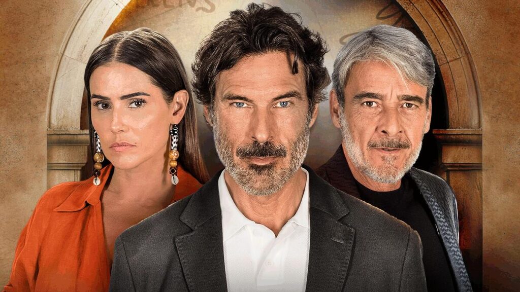 Em primeiro plano vê um homem meia idade com barba para fazer de camisa e terno preto. Ao fundo, do lado direito, vê-se Alexandre Borges, grisalho e barba semi cerrada. do outro lado, Deborah Secco aparece de vermelho com brincos compridos