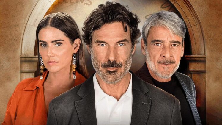 Em primeiro plano vê um homem meia idade com barba para fazer de camisa e terno preto. Ao fundo, do lado direito, vê-se Alexandre Borges, grisalho e barba semi cerrada. do outro lado, Deborah Secco aparece de vermelho com brincos compridos