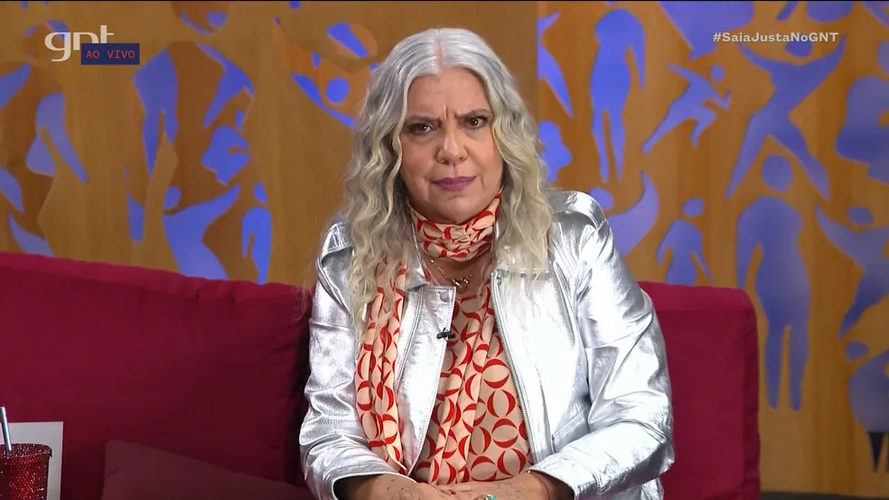 Astrid Fontenelle está sentada com casaco prateado e camisa laranja.