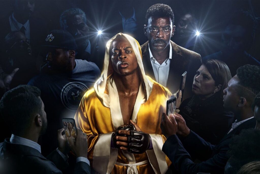A imagem mostra o lutador Anderson Silva vestido com robe amarelo dourado cercado de fotógrafos ofuscado por seu luz. Ao fundo se vê o ator Seu Jorge, tio de Anderson e apoio familiar.