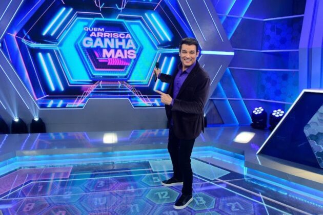 Celso Portiolli no centro do palco do Domingo Legal com a logomarca do Quem Arrisca Ganha Mais