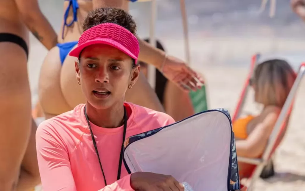 Raquel com viseira rosa na praia vendendo sanduíche