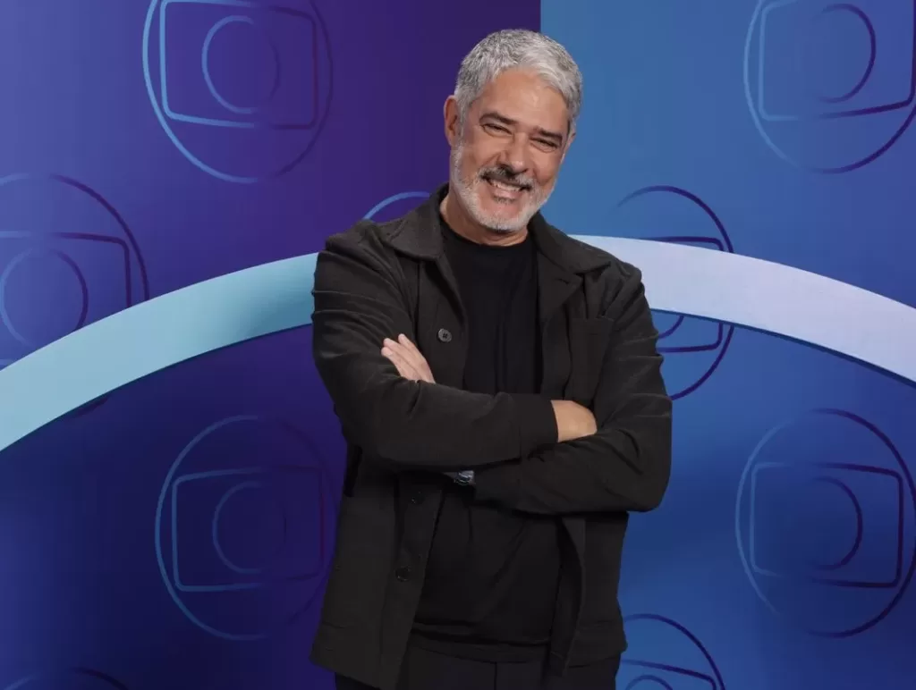 Willian Bonner de braços cruzados à frente da logo da Globo